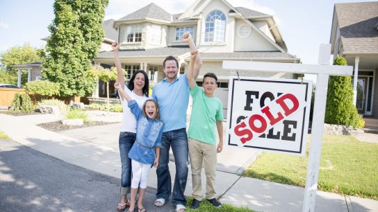A family of four are standing outside of the home they just sold. The sign in front of the house says, "for sale" and "sold." They are smiling, cheering and looking at the camera.