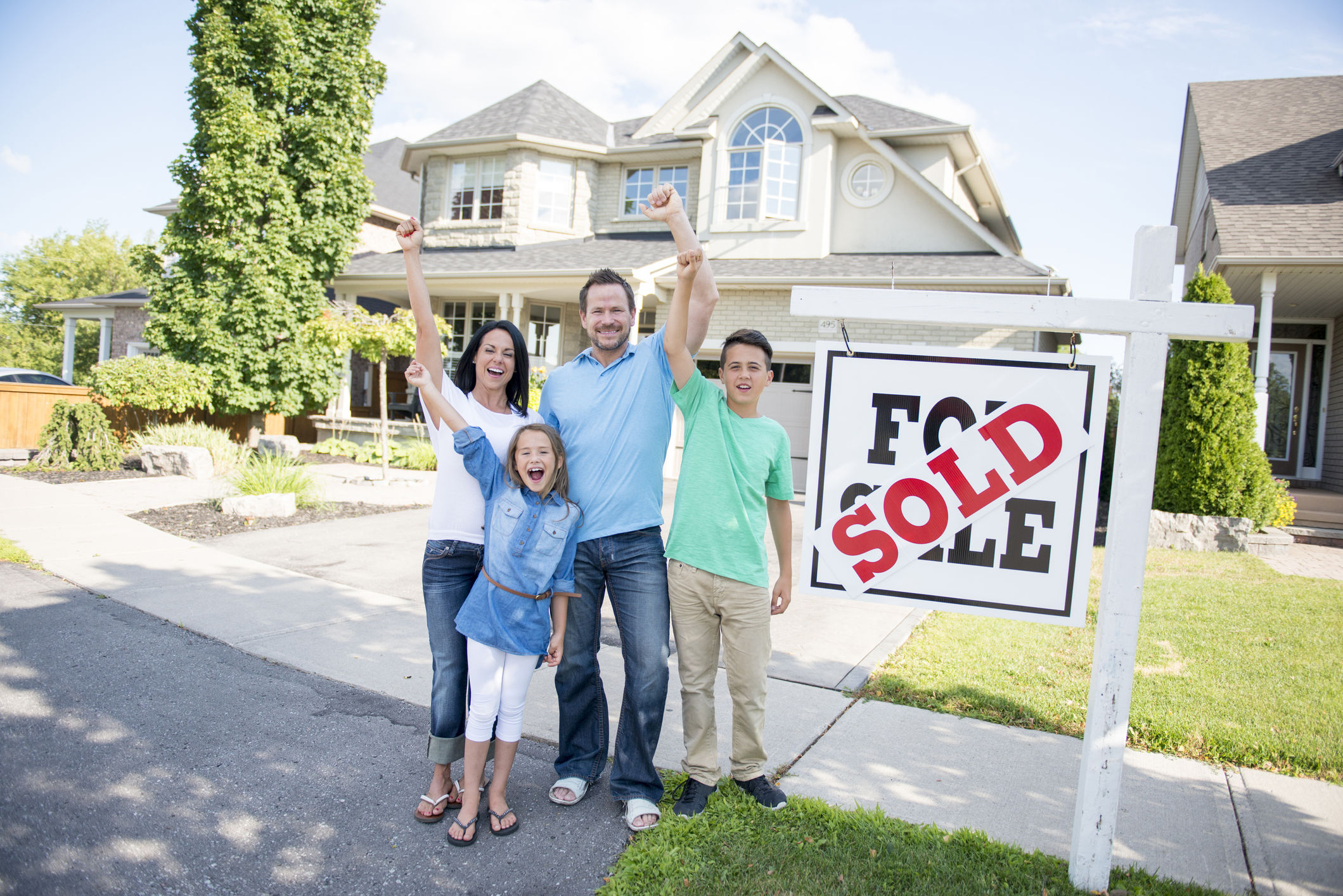 A family of four are standing outside of the home they just sold. The sign in front of the house says, "for sale" and "sold." They are smiling, cheering and looking at the camera.